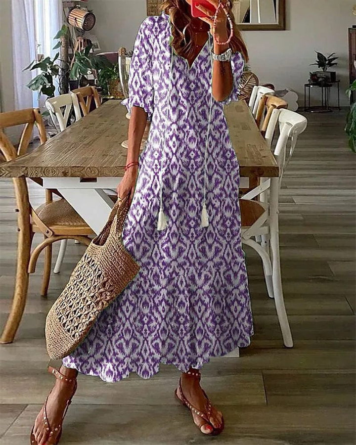 Three quarter view of model wearing a purple and white printed summer dress with a woven bag and sandals
