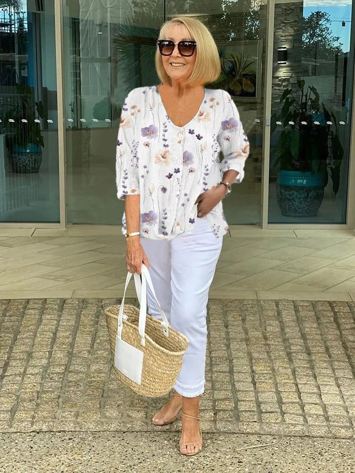 Full length view, model smiling, wearing a floral button-up blouse, white pants, sunglasses and holding a straw bag