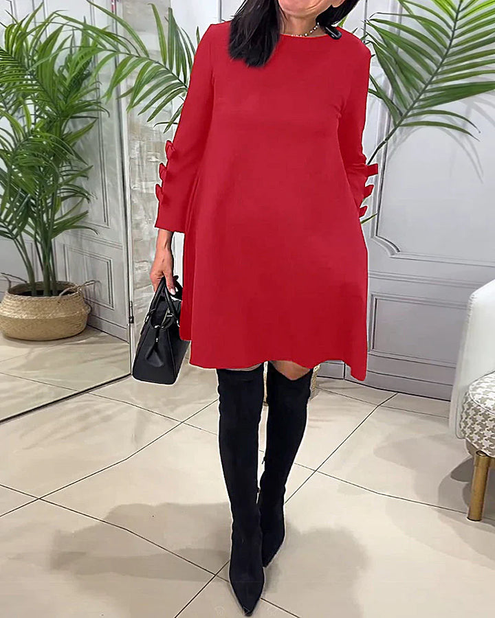 Front view worn by model, a red relaxed long-sleeve knee-length dress with bow details on the sleeves and handbag