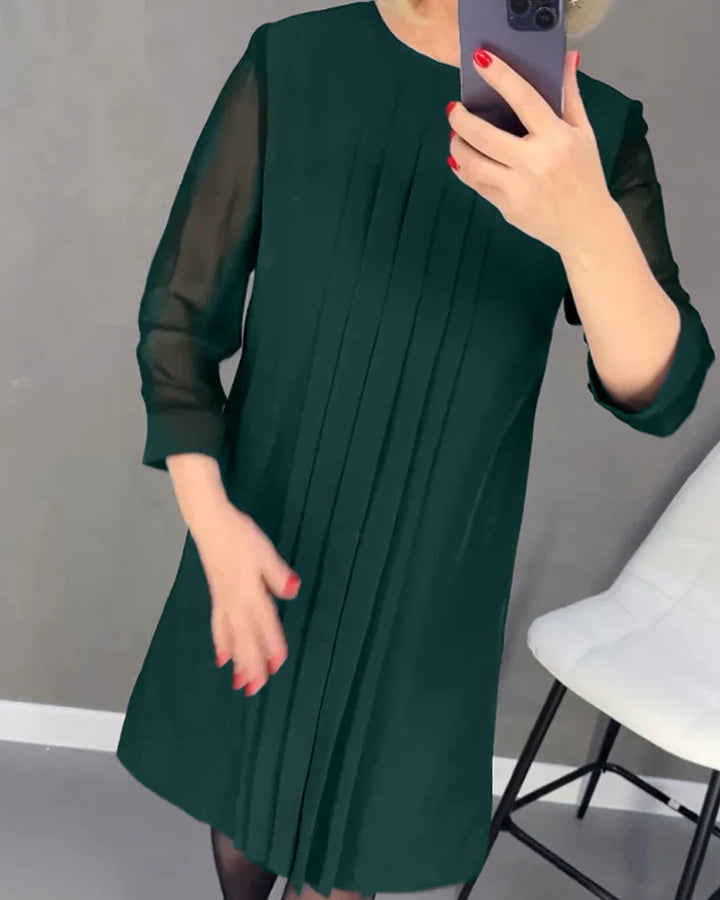 Front view, model in a dark green pleated knit dress with sheer sleeves, holding a phone to her face
