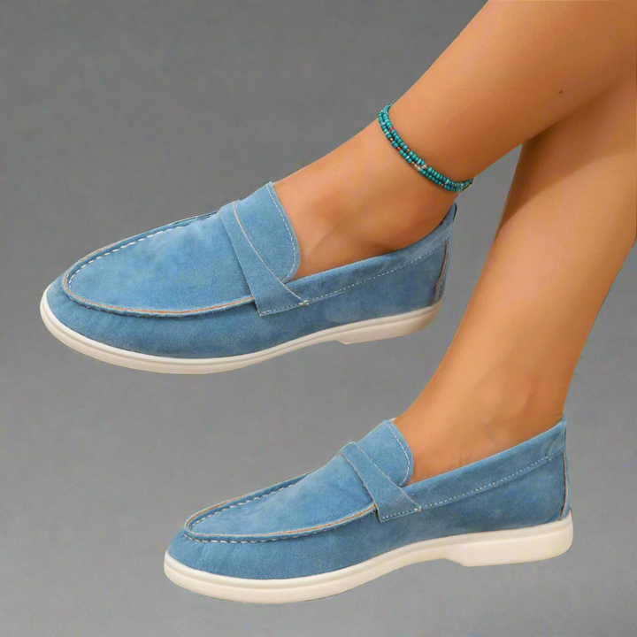 Side view of blue suede Ellis Elegant Flat Shoes with white soles and a turquoise beaded anklet
