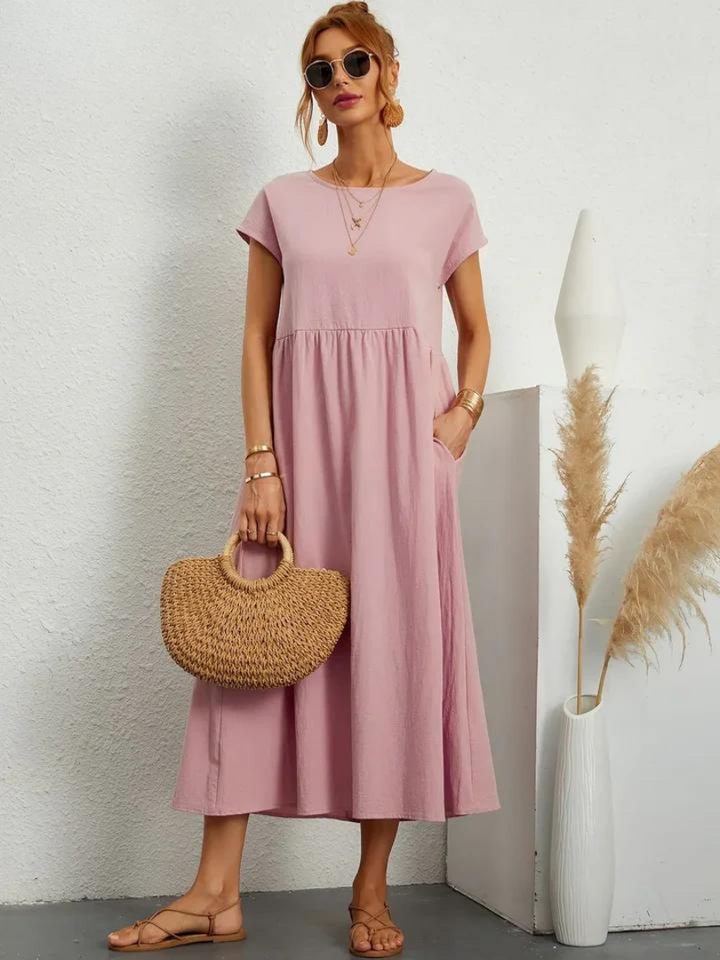 Front view, model wearing a pink loose summer dress with short sleeves, sunglasses and a woven handbag