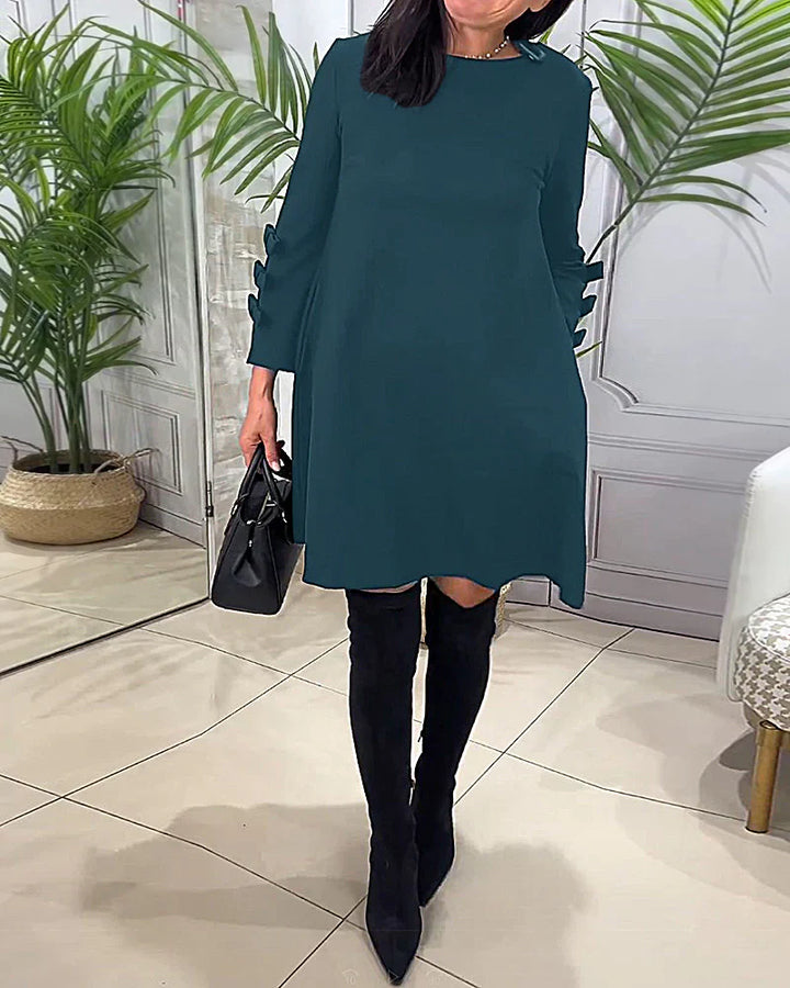 Front view worn by model, a blue relaxed long-sleeve knee-length dress with bow details on the sleeves and boots