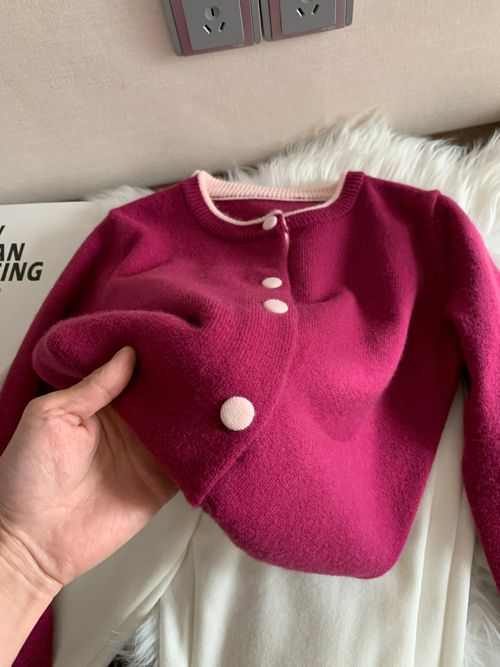 Front view of a hand holding a fuchsia knitted cardigan with light pink buttons and trim