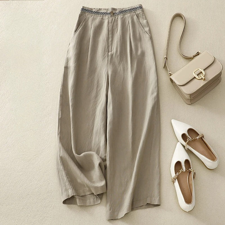 Flat lay of beige linen pants with a patterned waistband, styled with a handbag and white flats