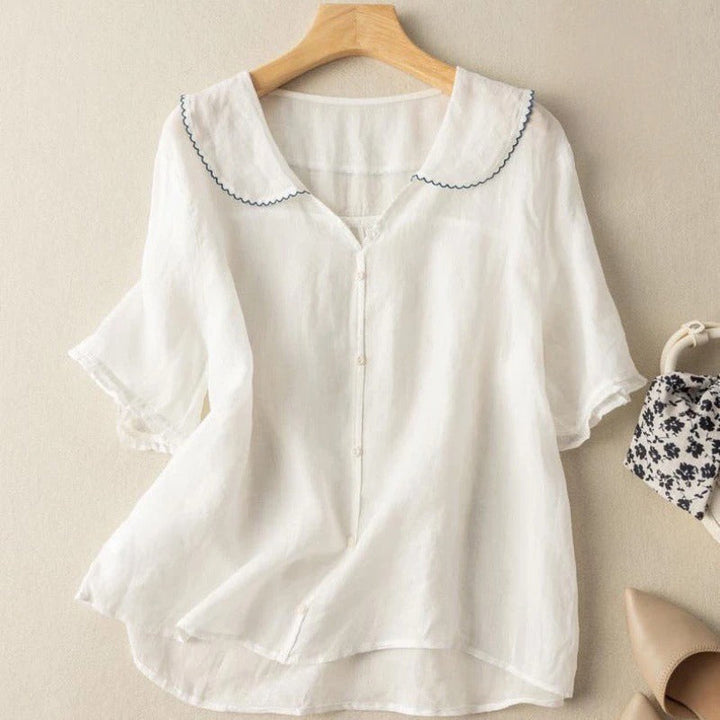 Front view of a white lightweight linen blouse with a blue scalloped collar and short sleeves on a hanger