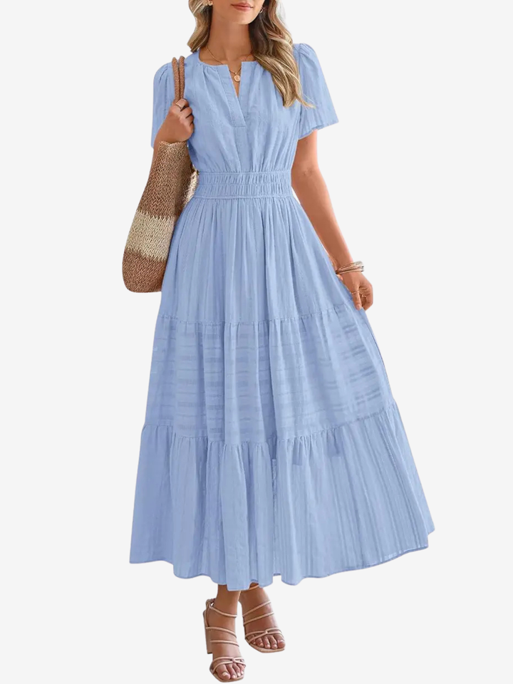 Front view of a model in a light blue tiered maxi dress with cinched waist, V neck, and short sleeves, holding a woven bag