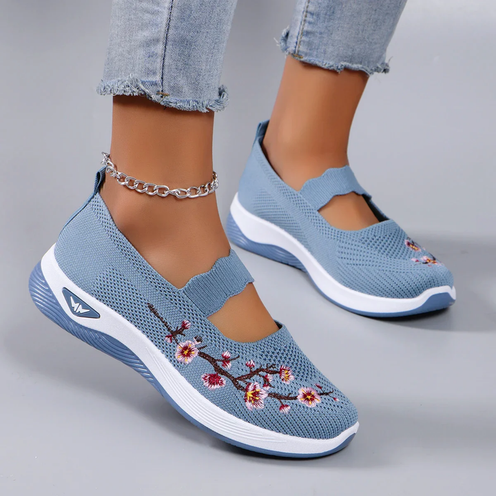 Three quarter view of model wearing blue breathable slip-on sneakers with pink floral embroidery and an ankle bracelet