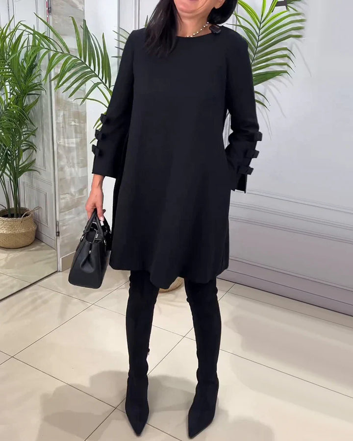 Front view worn by model, a black relaxed long-sleeve knee-length dress with bow details on the sleeves