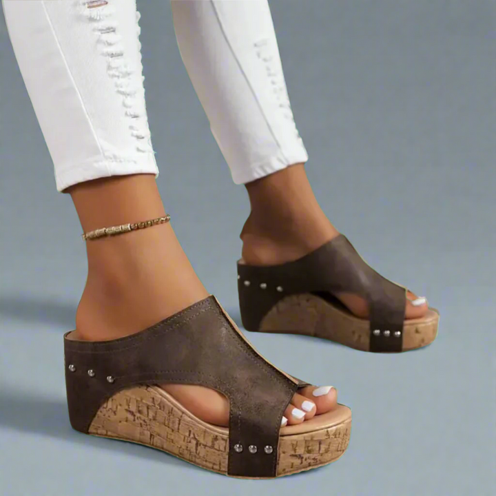 Worn by model, two feet in elegant summer wedge sandals with brown straps and cork-look soles, white jeans
