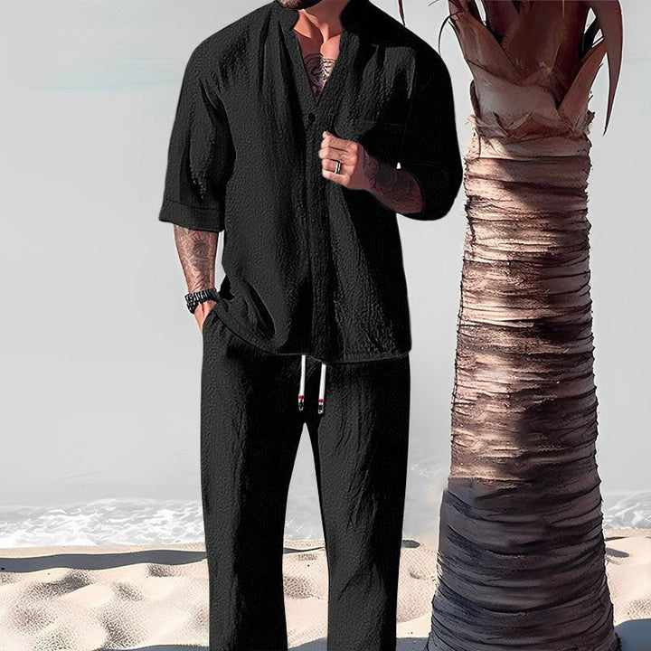 Front view of a model wearing a black cotton-linen V-neck top and trouser set on a beach next to a palm tree