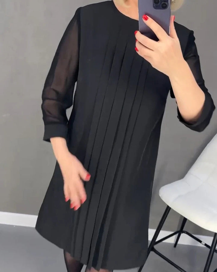 Front view, model wearing a black pleated knit dress, holding a phone to her chest, with a white chair beside her