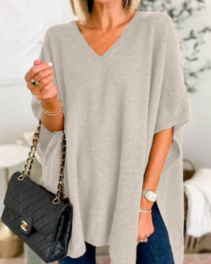Front view of model wearing the Erica oversized V-neck poncho sweater in light grey, holding a black quilted handbag