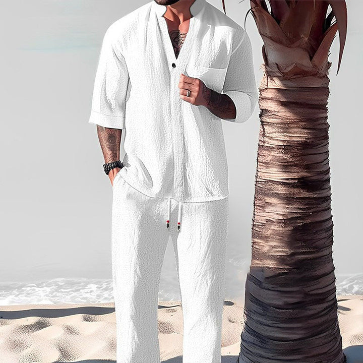 Front view of a model wearing a white cotton-linen V-neck top and trouser set on a beach next to a palm tree