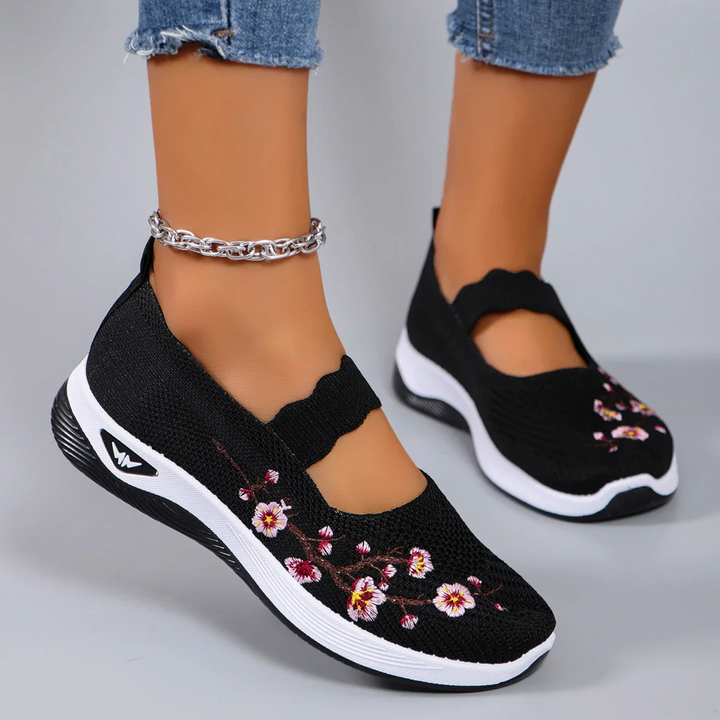 Close up of model wearing black breathable slip-on sneakers with pink floral embroidery and an ankle bracelet