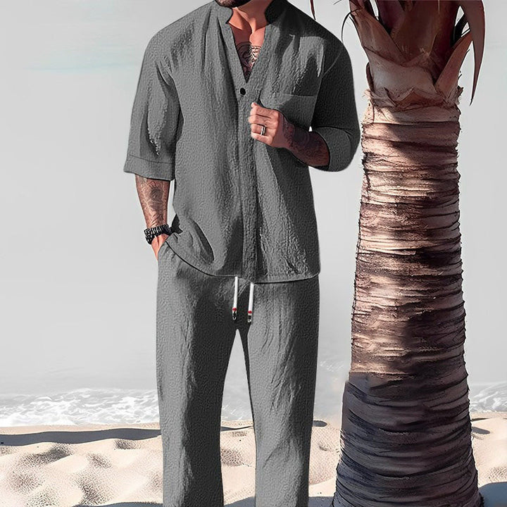 Front view of a model wearing a grey cotton-linen V-neck top and trouser set on a beach next to a palm tree