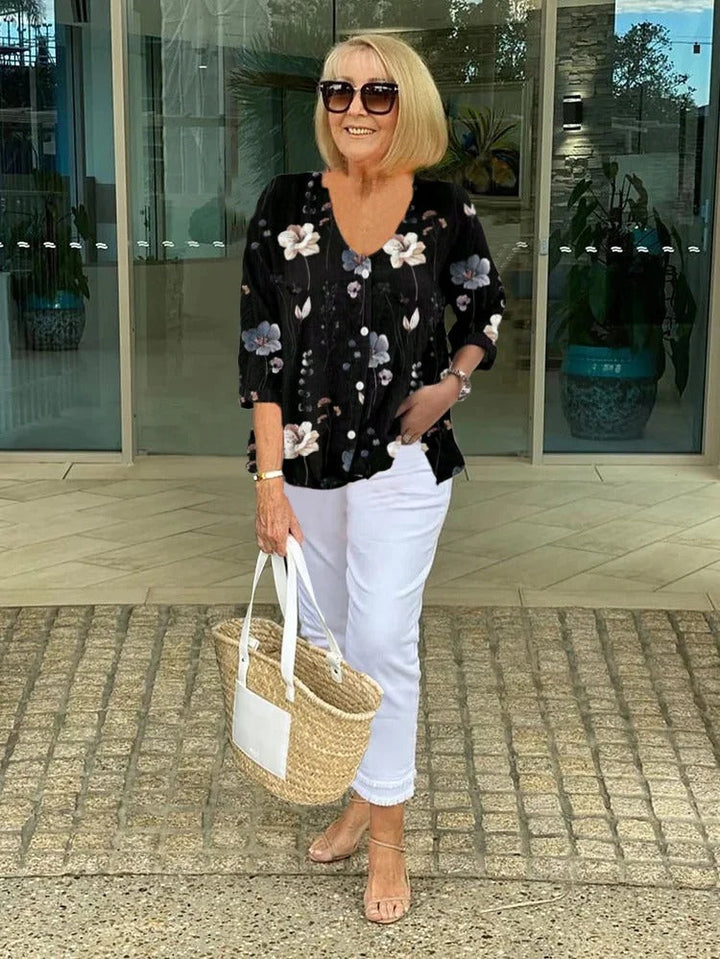 Model smiling, wearing a floral button-up blouse, white capri pants, sunglasses and holding a straw bag