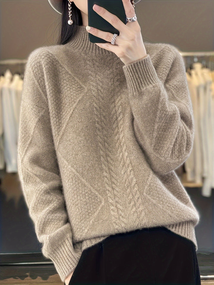 Front view of a model wearing a light brown cable knit turtleneck sweater with diamond patterns, holding a phone