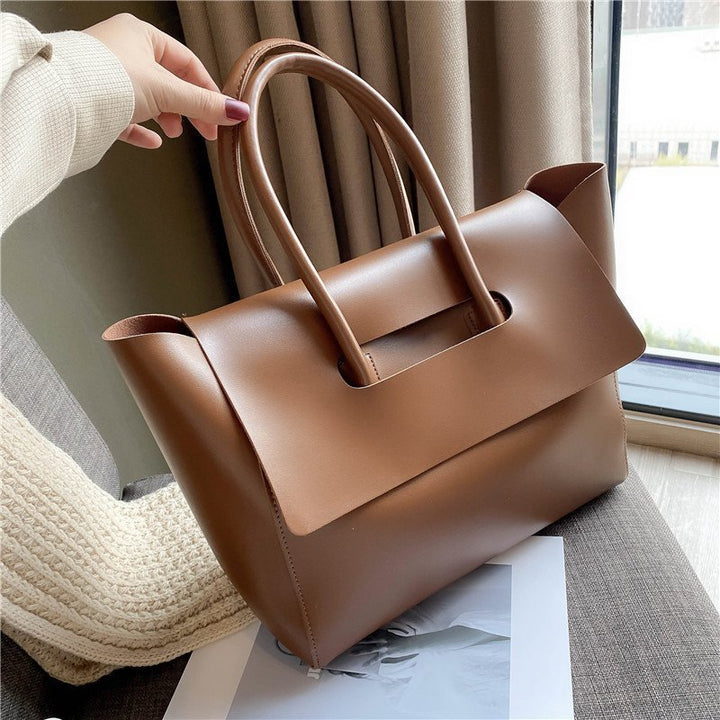 Three quarter view of a hand holding the top handle of a tan Geneva vegan leather handbag