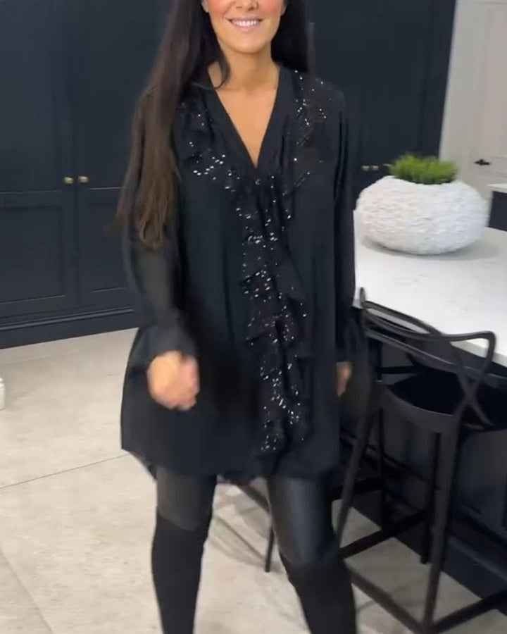 Front view of a woman wearing a black ruffled blouse with sequin detail and black pants, smiling at the camera