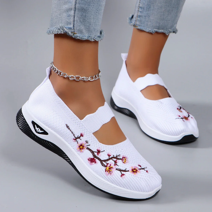 Close up of model wearing white breathable slip-on sneakers with pink floral embroidery and an ankle bracelet