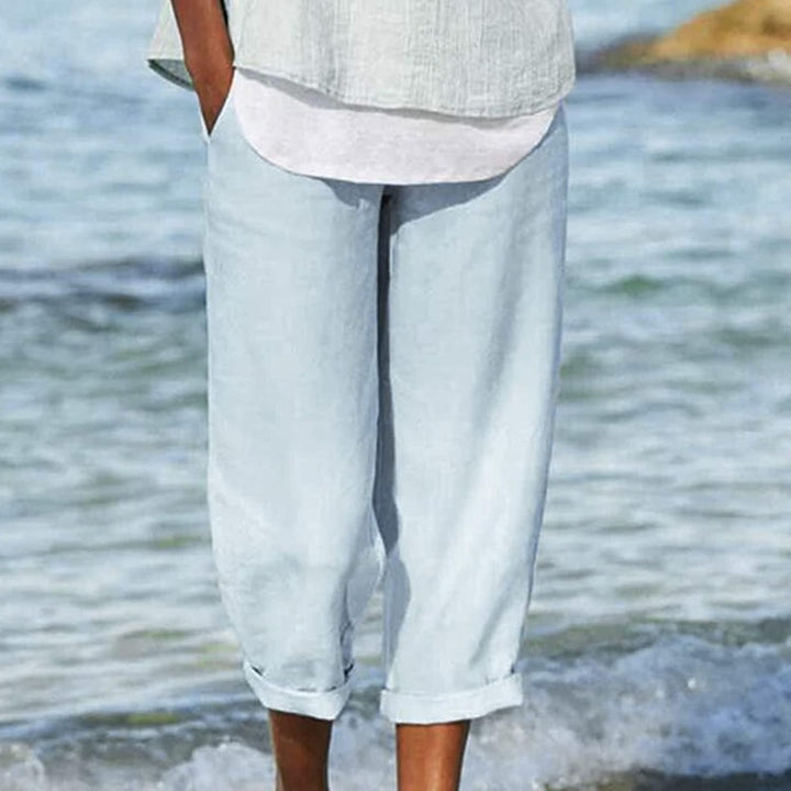 Front view of model wearing breezy cropped pants in light blue, standing on a beach with water lapping at her feet