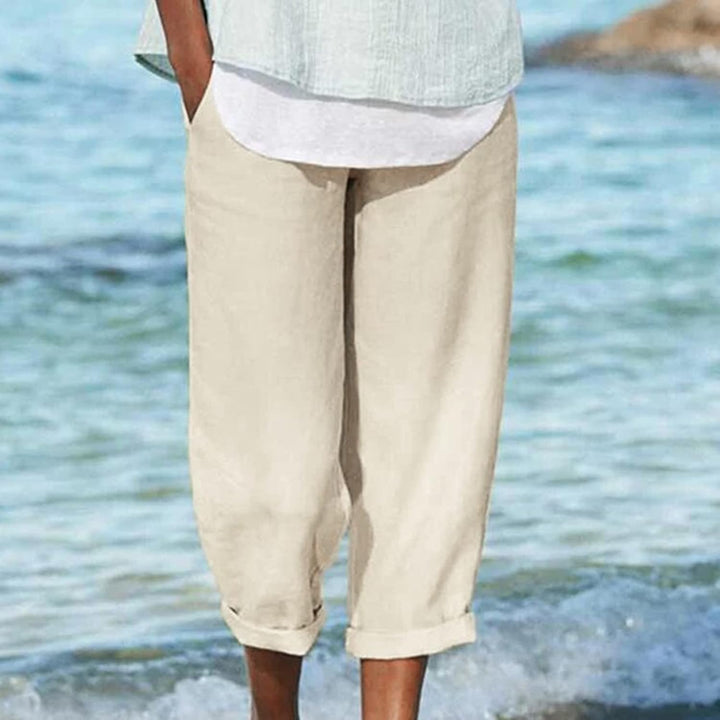 Back view of model wearing breezy cropped pants in white, standing on a beach with water lapping at her feet