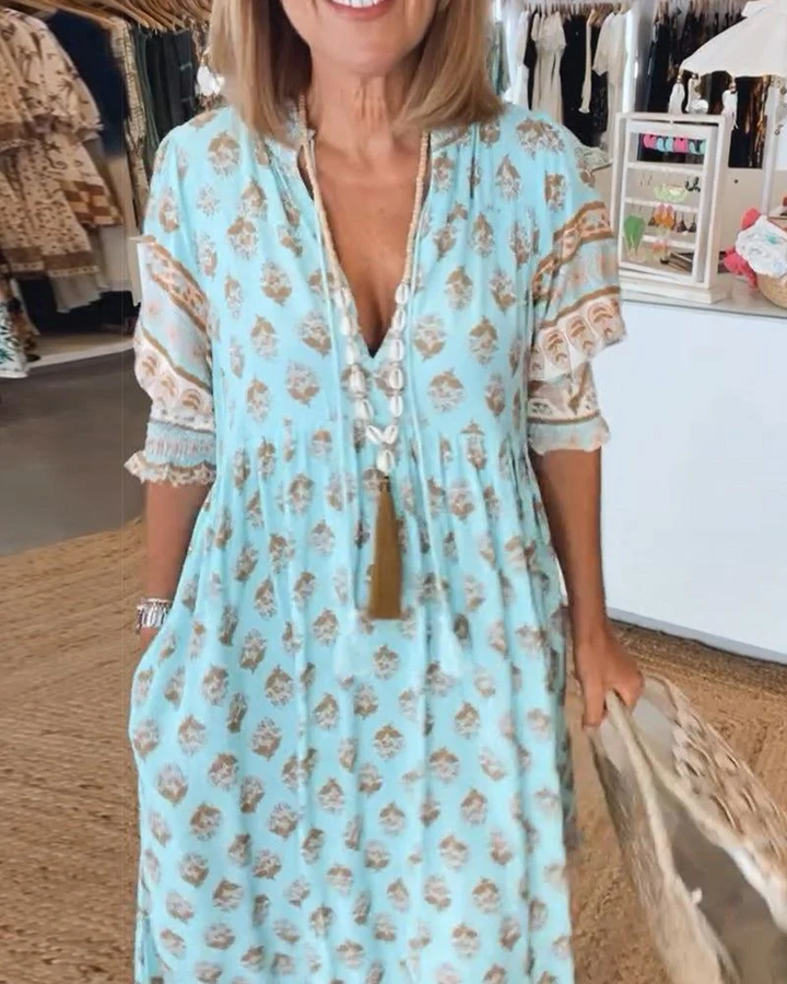Close up of a woman wearing a light blue casual V-neck dress with a gold floral print and a shell necklace