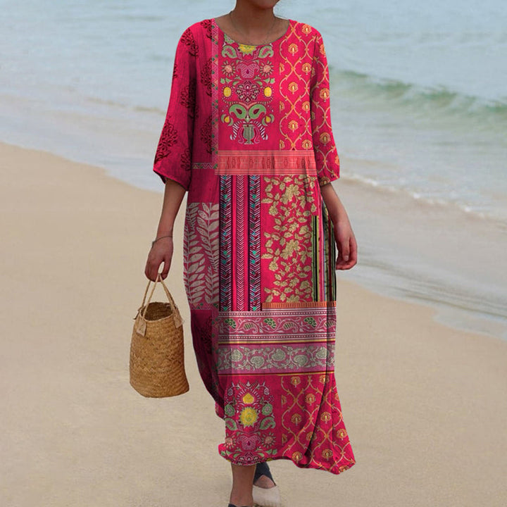 Front view of a woman wearing a flowing gown with a vibrant bohemian print, holding a woven bag on a sandy beach
