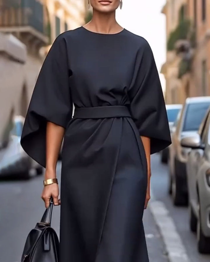 Three quarter view of a model in a black kimono sleeve midi dress with an elegant belted waist and handbag