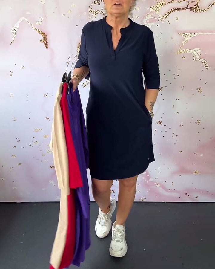 Front view of a woman wearing a navy V-neck dress, holding other colours of the dress on hangers