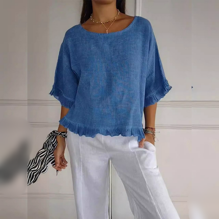 Front view, Zelda casual elegant blouse in blue with ruffle detail, worn by model with white pants