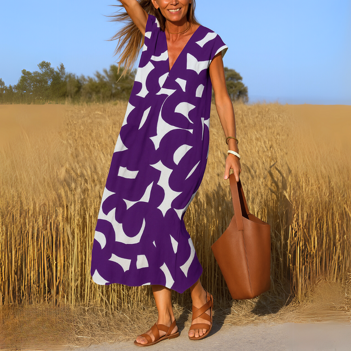 Front view of a woman wearing a purple and white patterned flowing V-neck boho dress with a brown bag