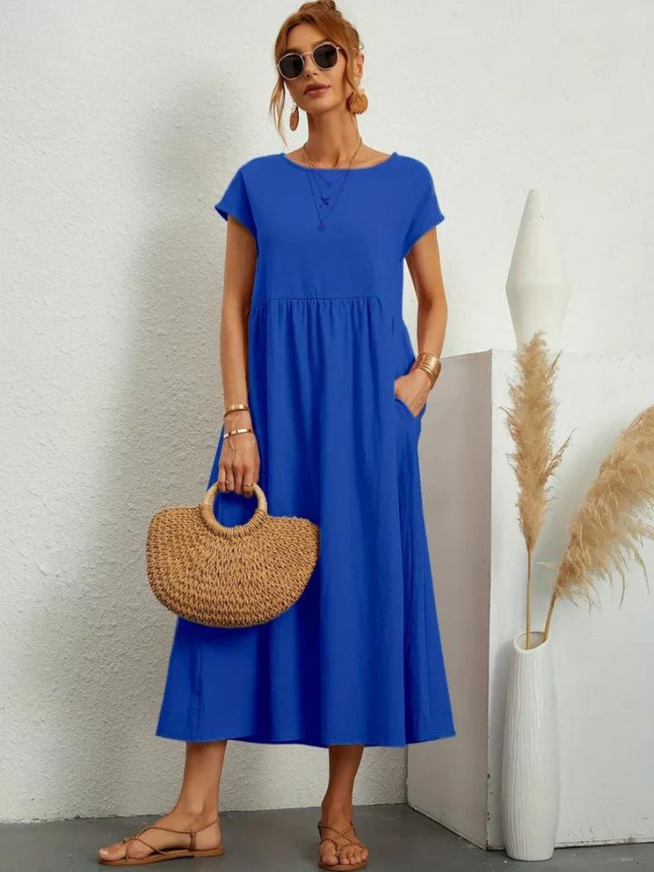 Front view, model wearing a blue loose summer dress with short sleeves, sunglasses and a woven handbag