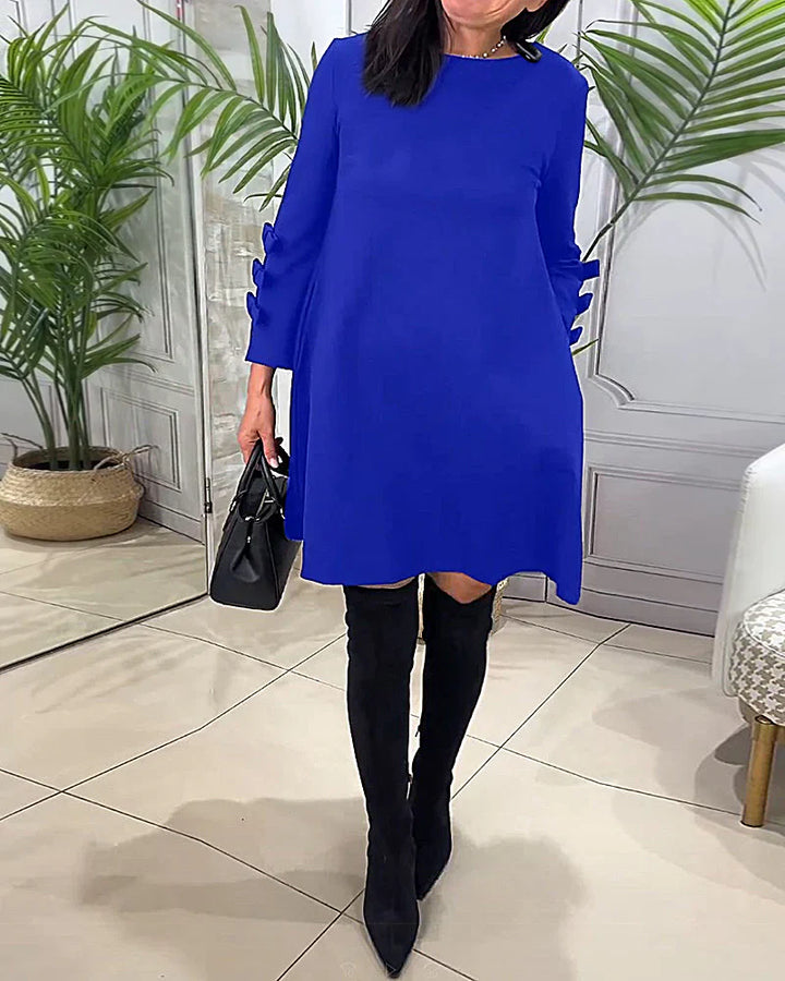 Front view worn by model, a blue relaxed long-sleeve knee-length dress with bow details on the sleeves and handbag