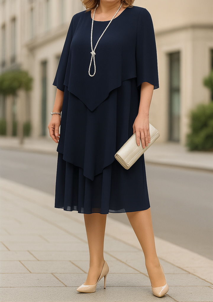 Three quarter front view of a woman wearing a navy blue elegant dress with a long pearl necklace and clutch bag