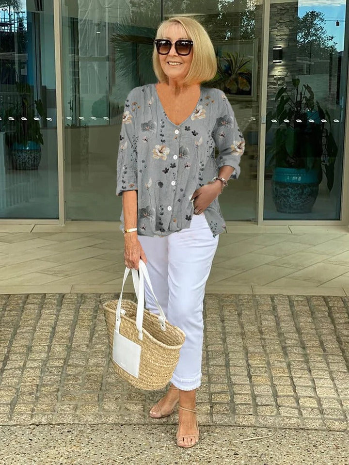 Model wearing a floral button-up blouse, white capri pants, sunglasses and holding a straw bag