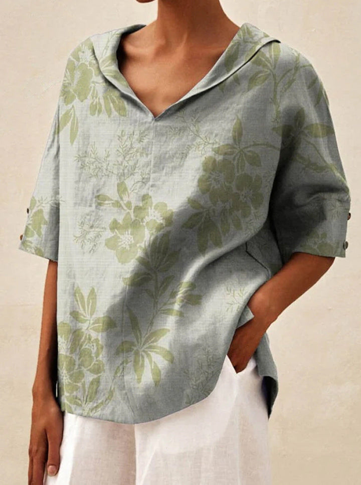 Close up front view of a model wearing a grey V-neck floral top with green flowers, hand in pocket