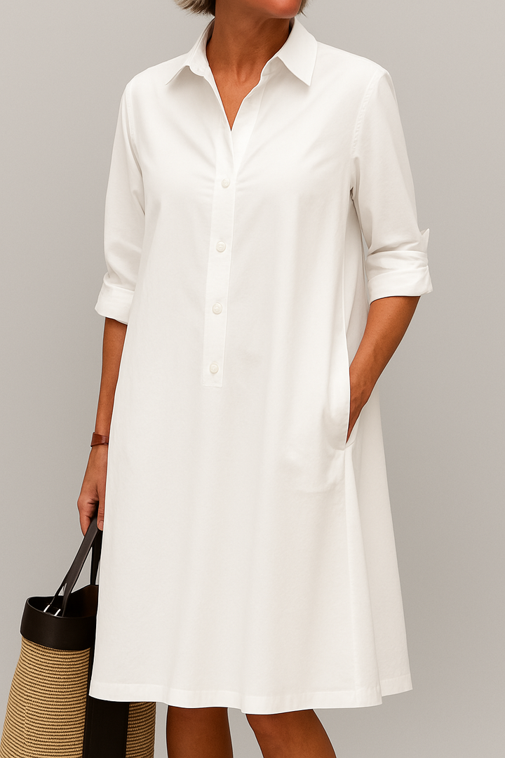 Front view, model wearing a white button-down shirt dress with rolled sleeves, one hand in a pocket, holding a straw bag
