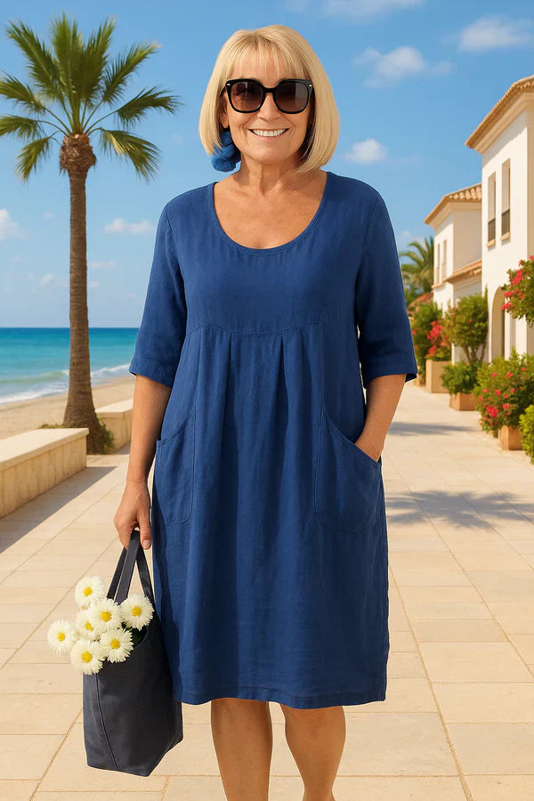 Front view of a woman in a blue linen summer dress with pockets, smiling with a hand in a pocket