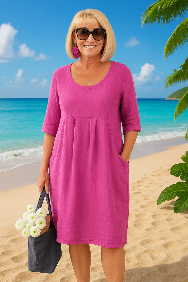 Front view of a smiling woman in a pink linen summer dress with pockets, holding a tote bag with flowers
