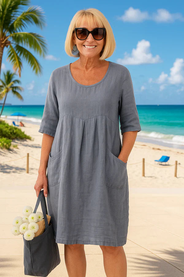 Front view of a smiling woman in a grey linen summer dress with pockets, holding a bag of flowers at the beach