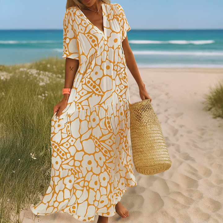 Front view, model in a white and yellow patterned summer dress holding a straw bag on a sandy path