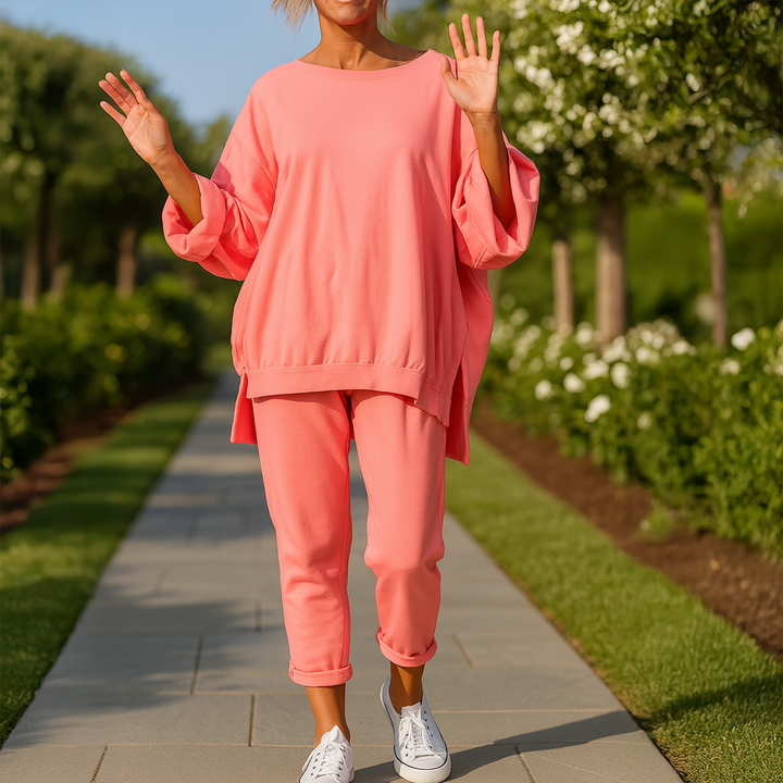 Front view of a model in a coral loose sweatshirt and trousers set with white sneakers on a garden path