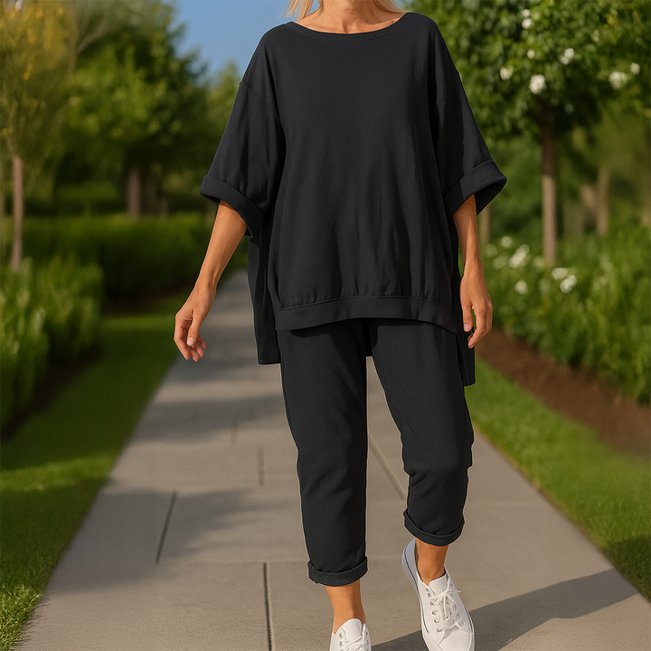 Front view of a model wearing a black loose sweatshirt and trousers set with white sneakers on a garden path
