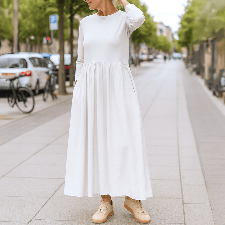 Front view of a woman wearing a white flowy knitted midi dress with long sleeves and pockets on a footpath