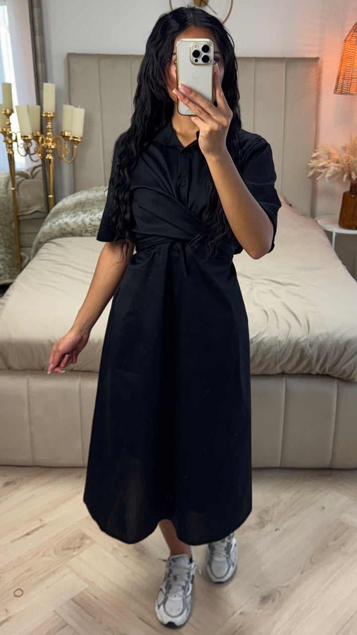 Front view, worn by model, a black cross tie belt blouse dress in soft cotton blend with a phone covering her face