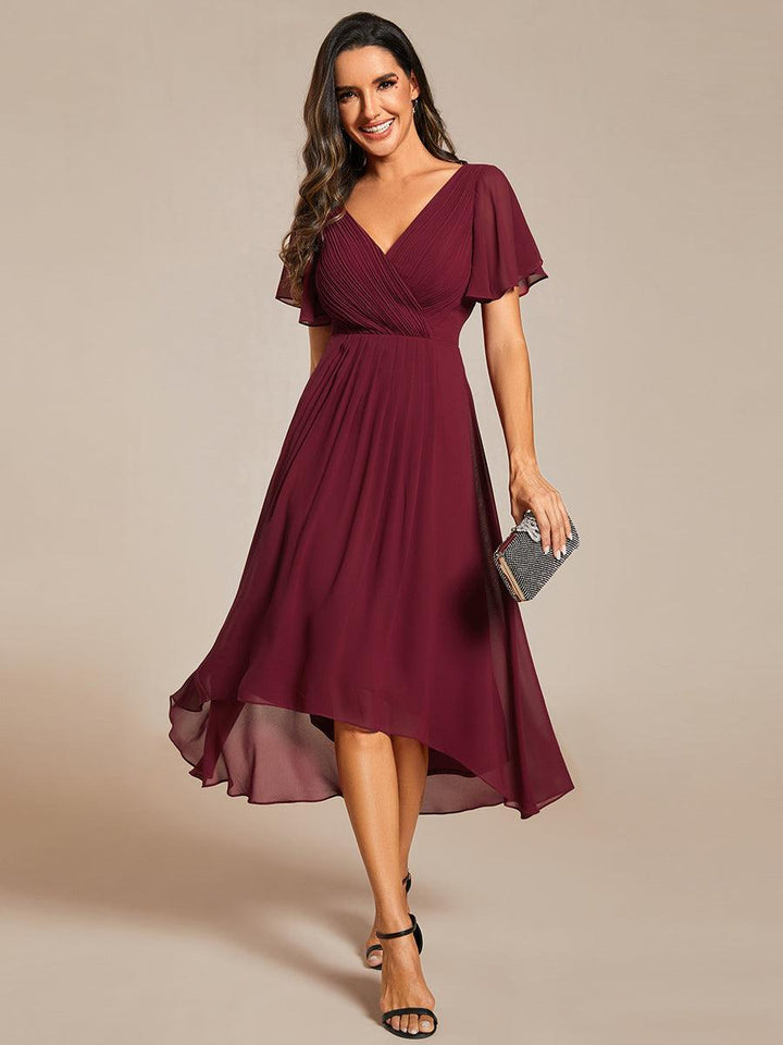 Front view, model in a rust chiffon V neck evening dress with flutter sleeves and high low hem, holding a clutch