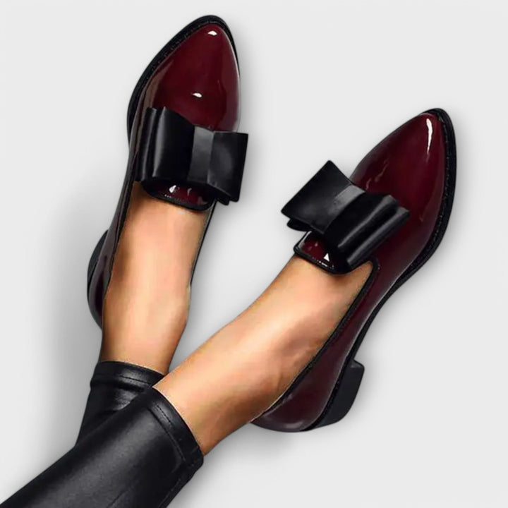 Worn by model, a pair of shiny burgundy loafers with black bows on the vamp, styled with black trousers