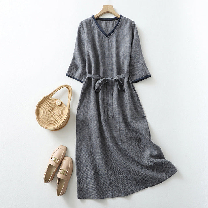 Front view of a grey V-neck linen summer dress with a self-tie belt, displayed with a straw bag and loafers